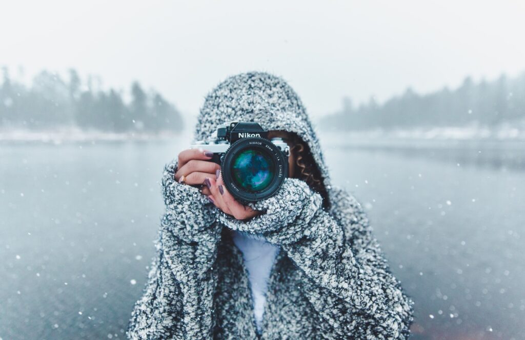 A person wrapped in a thick, cozy sweater holds a camera up to their face, capturing a snowy scene by the water. The snowfall gently frames the serene background of trees and water. This image emphasizes the importance of packing warm, layered clothing and capturing winter memories.