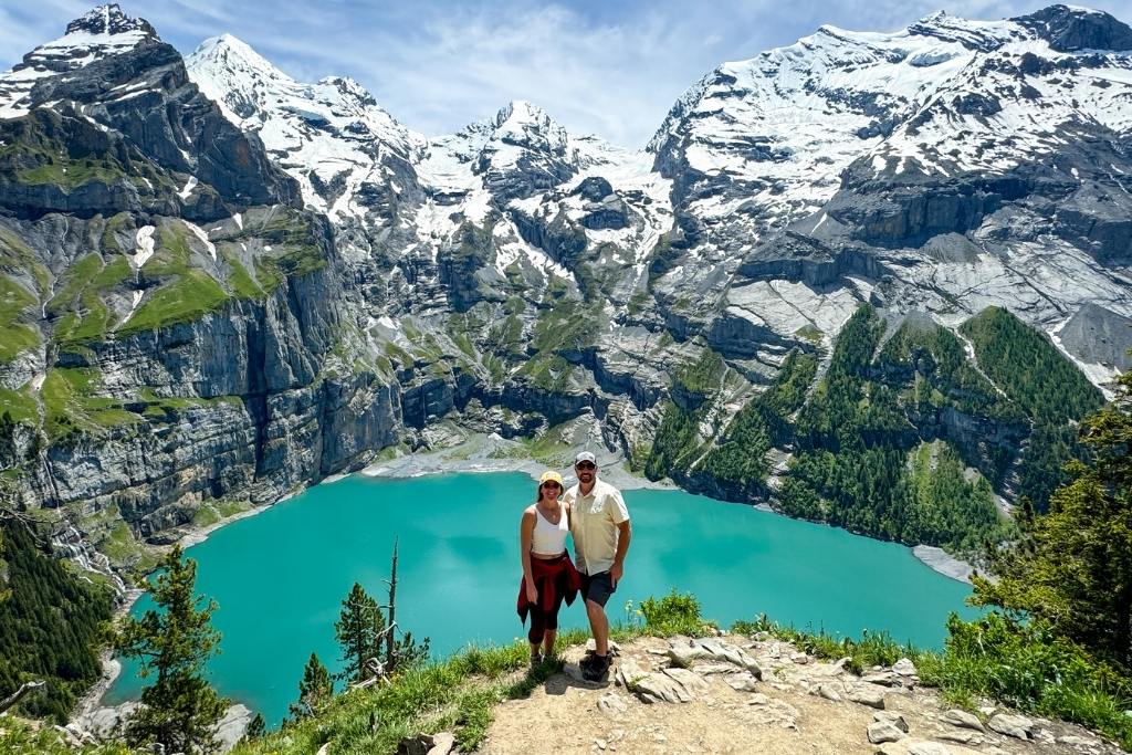 Kate from Kate's Crossing Blog and her husband standing on a scenic mountain trail overlooking a vibrant turquoise alpine lake in Switzerland. Snow-capped peaks and lush greenery surround the lake, creating a stunning natural backdrop. Both are dressed in hiking attire, smiling and enjoying the breathtaking views.