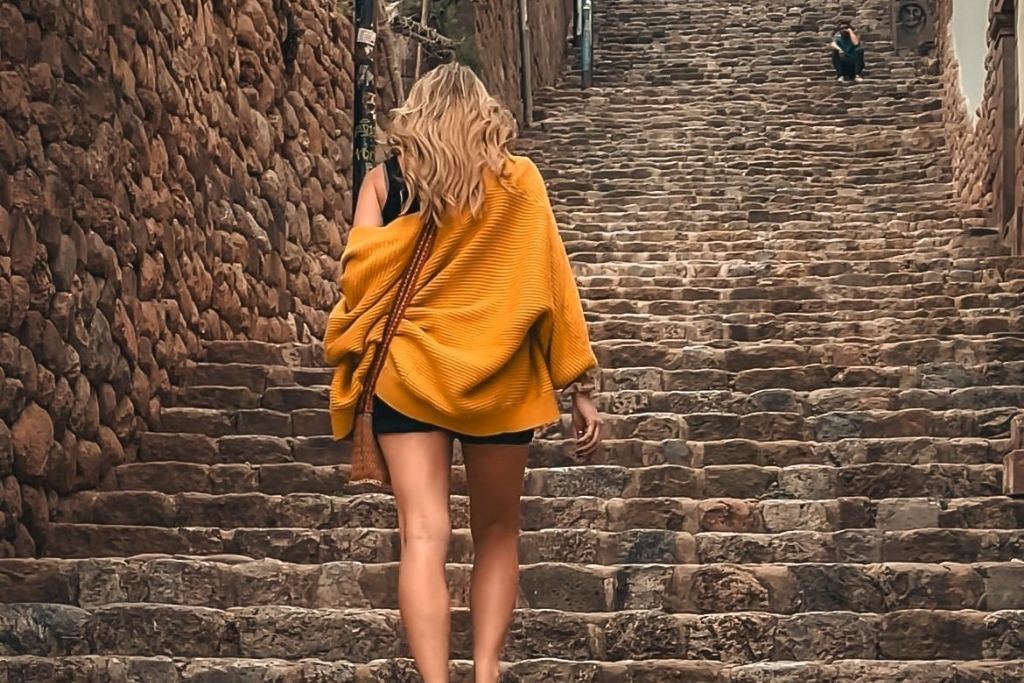 Kate wrapped in a mustard yellow shawl walks up a steep, narrow stone staircase in Cusco, Peru. The path is flanked by rustic stone walls and traditional buildings, capturing the city's historic charm and character.