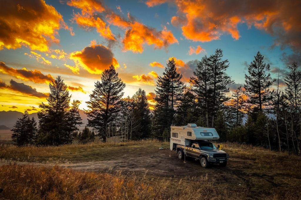 A scenic campsite in Teton National Park featuring a truck camper parked amidst tall pine trees during a vibrant sunset. The sky glows with dramatic orange and pink hues, contrasting with the silhouette of the trees and surrounding wilderness.