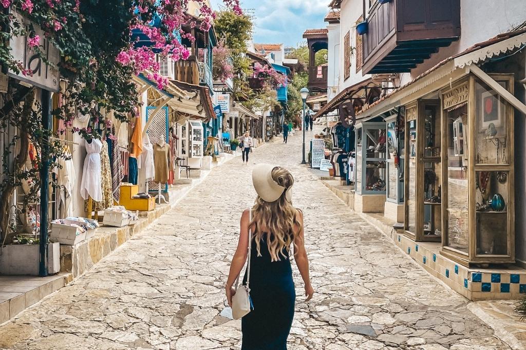 A charming street in Kas, Turkey, lined with vibrant shops, colorful flowers, and hanging bougainvillea. Kate is wearing a black dress and wide-brimmed hat walks along the cobblestone path, enjoying the quaint and picturesque surroundings.