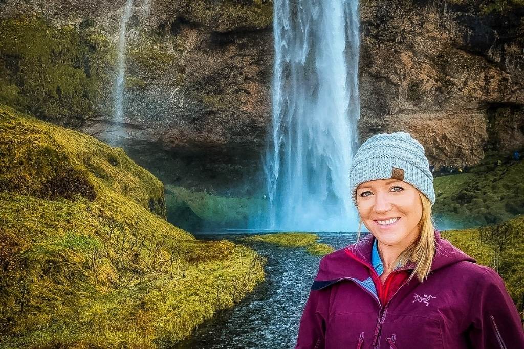 Kate in a purple jacket and gray beanie stands in front of the majestic Seljalandsfoss waterfall in Iceland. The cascading water is surrounded by lush green moss and a flowing stream, creating a serene and breathtaking scene.