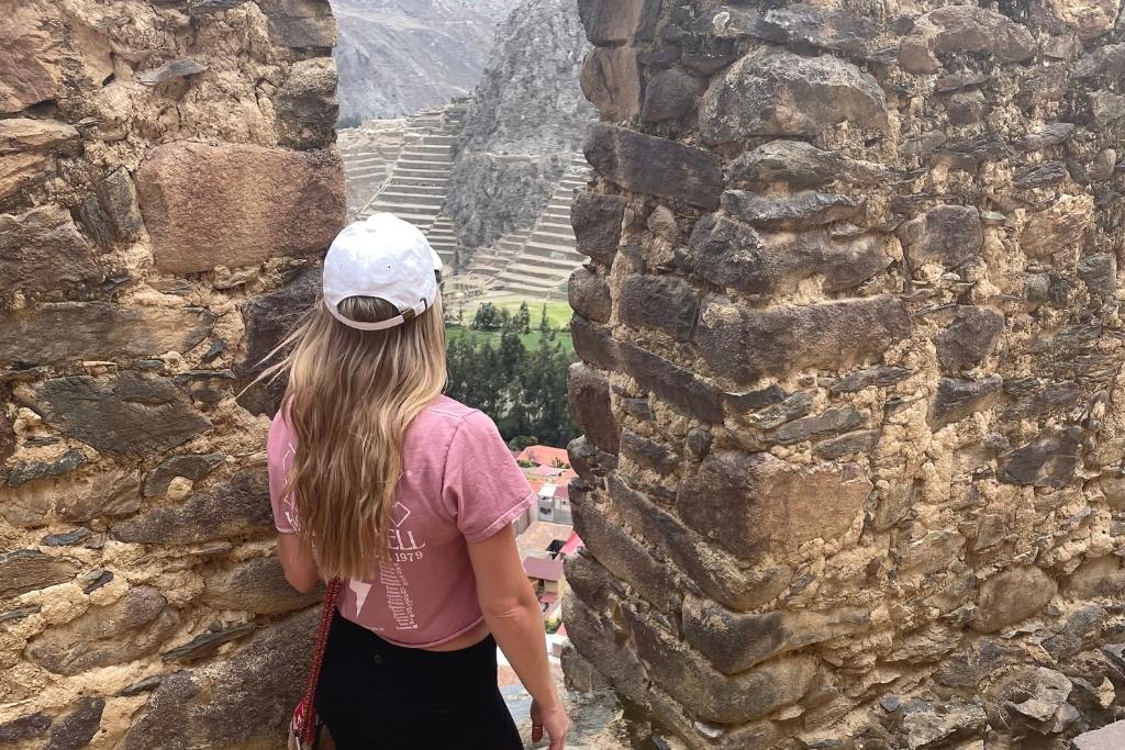 Kate wearing a pink T-shirt, black leggings, and a white cap stands at a stone doorway overlooking the ancient terraced landscape of Ollantaytambo in Peru. The view through the doorway showcases the iconic Inca ruins and lush green valleys below.