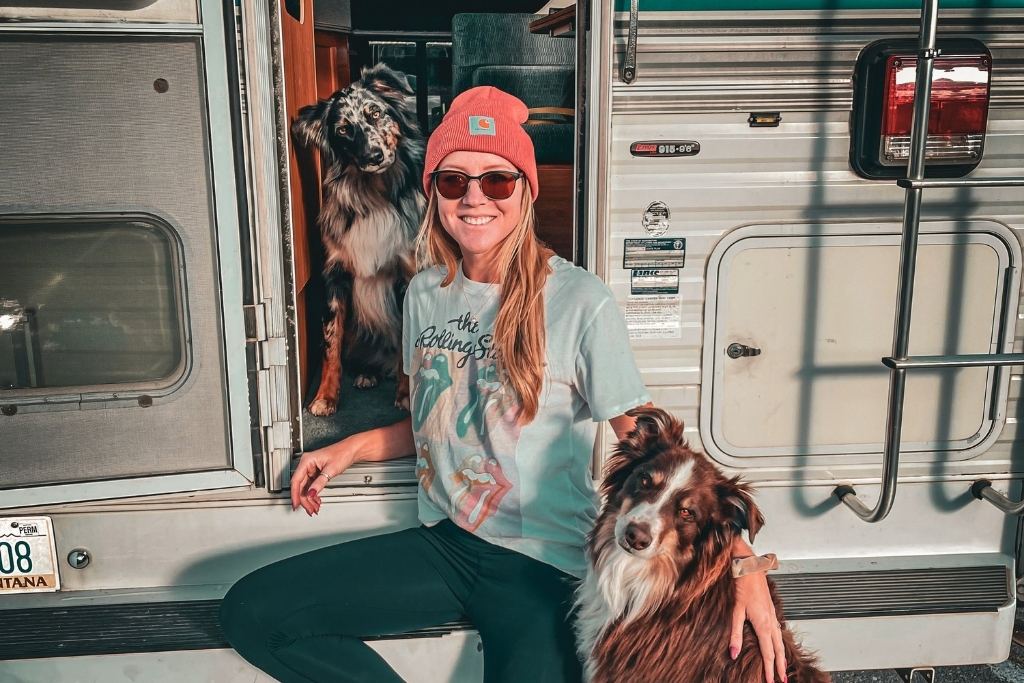 Kate wearing a Rolling Stones T-shirt, dark leggings, and an orange beanie sits on the step of a campervan, smiling with two dogs—one brown and white, and the other black and tan. The camper door is open, revealing the cozy interior. The scene suggests an adventurous road trip with pets.