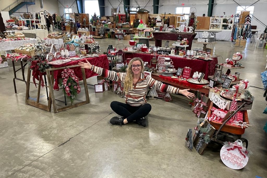 Kate sits cross-legged on the floor at a craft show, smiling and stretching her arms out to showcase a variety of festive holiday-themed crafts. The display features tables adorned with red tablecloths, wreaths, mugs, signs, and other Christmas decorations, set in a spacious indoor venue filled with other vendor booths in the background.