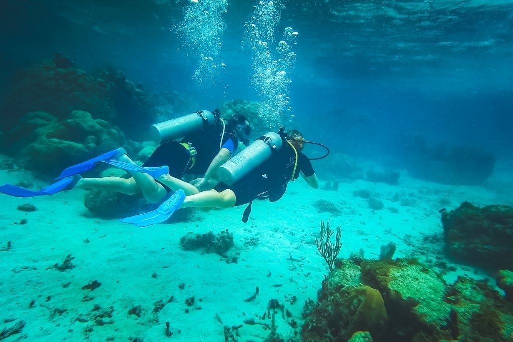 Kate from Kate's Crossing Blog and her husband scuba diving in the clear waters of Belize, surrounded by vibrant coral reefs and marine life. They are wearing full scuba gear, including tanks and fins, as they explore the underwater world. The serene blue hues and reef details highlight the beauty of the tropical marine environment.