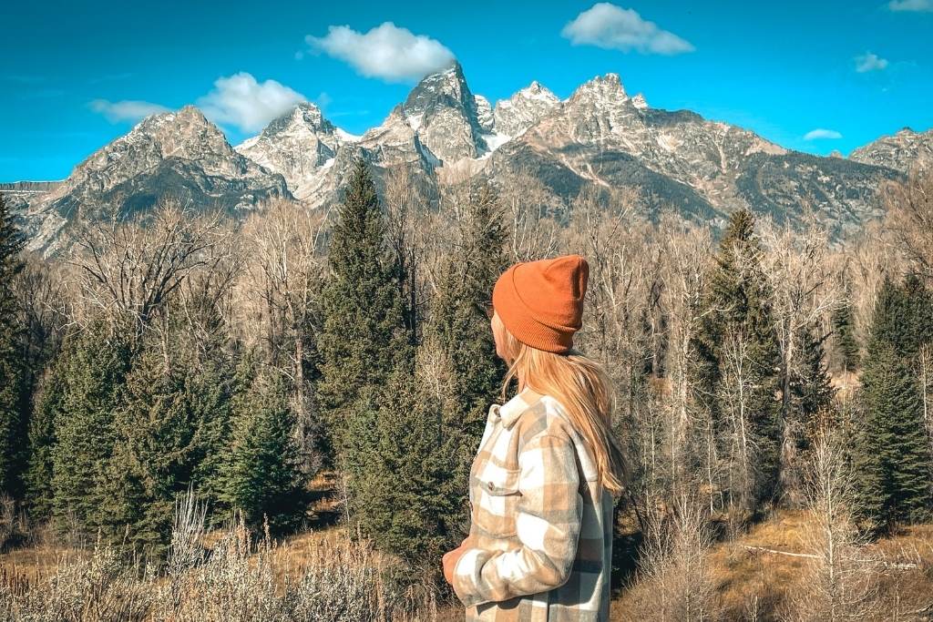 Kate from Kate's Crossing Blog gazing at the majestic peaks of the Teton Range in Grand Teton National Park. She is wearing a warm plaid jacket and an orange beanie, surrounded by a serene forest of evergreens and bare trees under a bright blue sky. The stunning mountain landscape provides a breathtaking backdrop to the tranquil moment.