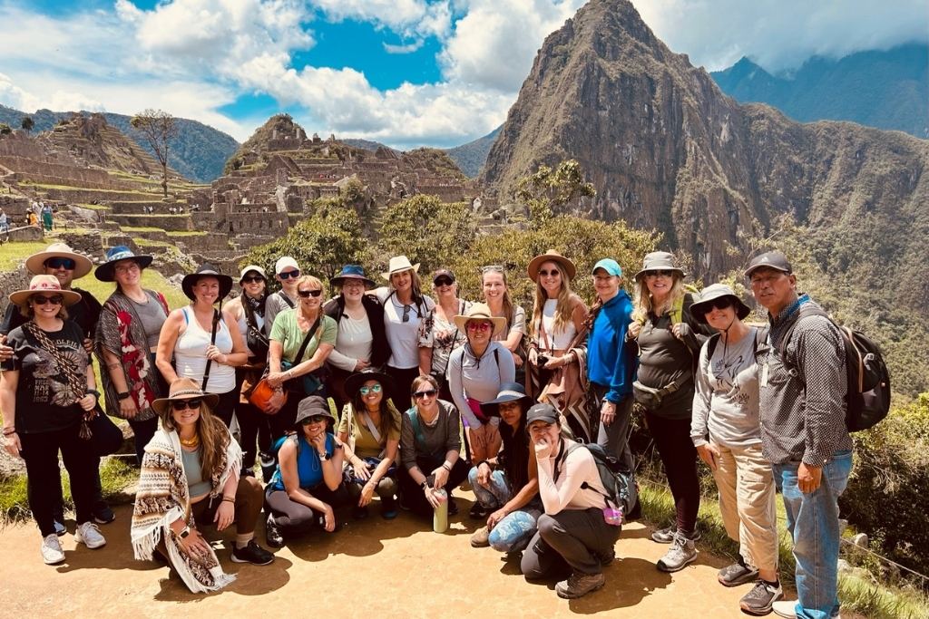 A group of travelers posing together at the iconic Machu Picchu site in Peru. They are surrounded by lush green mountains and the ancient Inca ruins under a bright blue sky. The group, dressed in hiking gear and sun hats, radiates excitement and camaraderie in this unforgettable adventure setting.