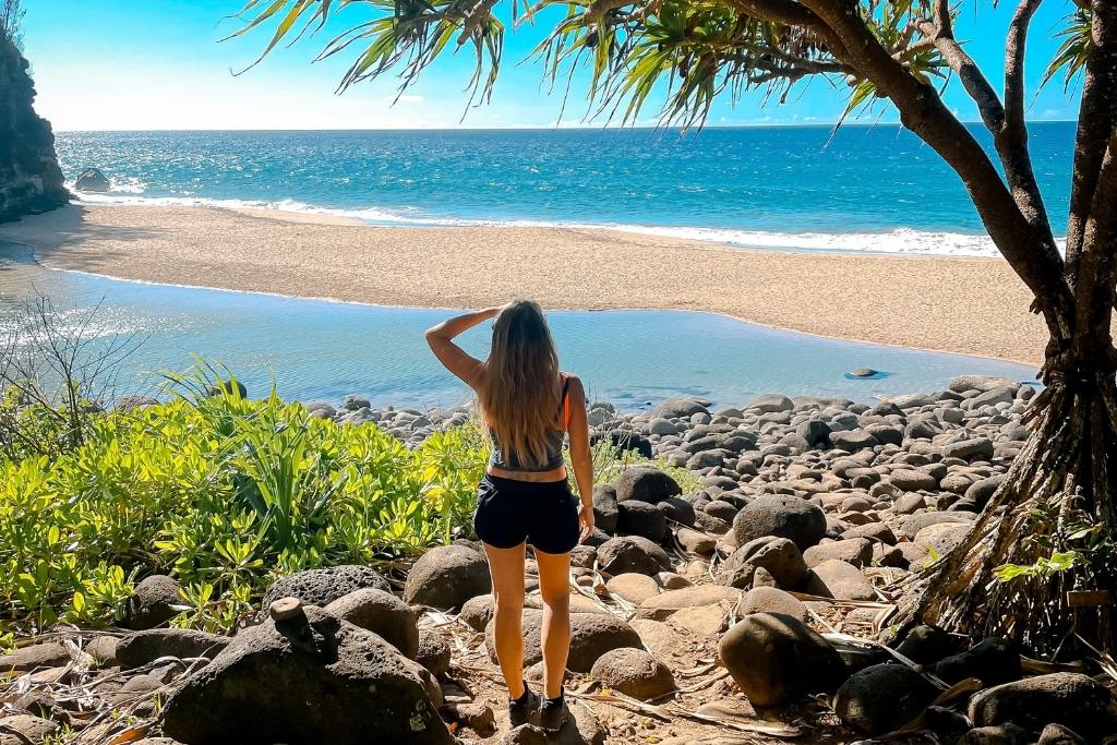 Kate stands on a rocky trail surrounded by lush greenery, gazing out at a serene beach and the vast ocean in Kauai, Hawaii. The golden sand, shimmering blue waters, and tropical foliage create a picturesque, tranquil scene under the bright sunlight. A perfect depiction of nature's beauty and peaceful solitude.