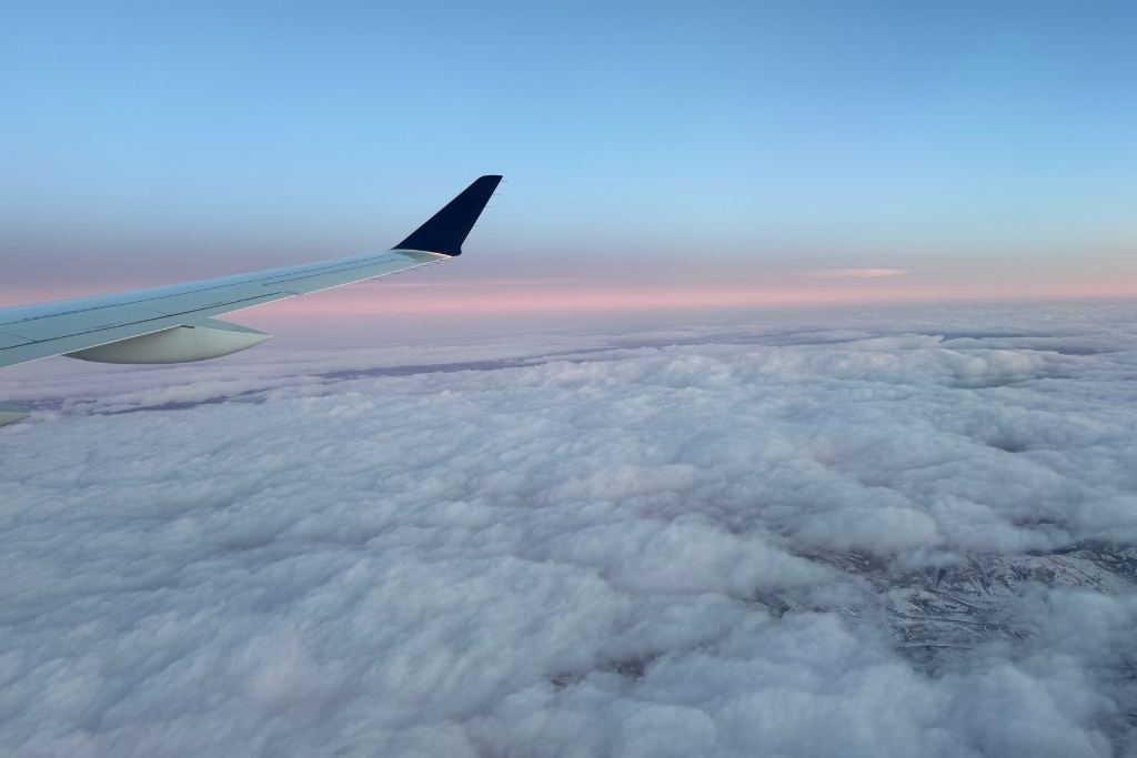 A serene view from an airplane window showing a vast expanse of fluffy white clouds stretching across the horizon, with the aircraft wing in the foreground. The sky above transitions from soft pastel pinks to a gentle blue, creating a tranquil and dreamy atmosphere.