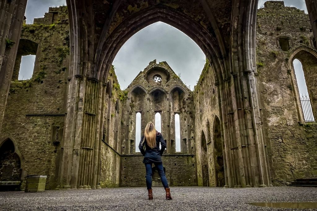 Kate stands in the middle of an ancient stone cathedral ruin in Ireland, surrounded by towering arches and weathered stone walls covered with patches of moss. The overcast sky adds a dramatic touch to the historic and gothic ambiance of the site.