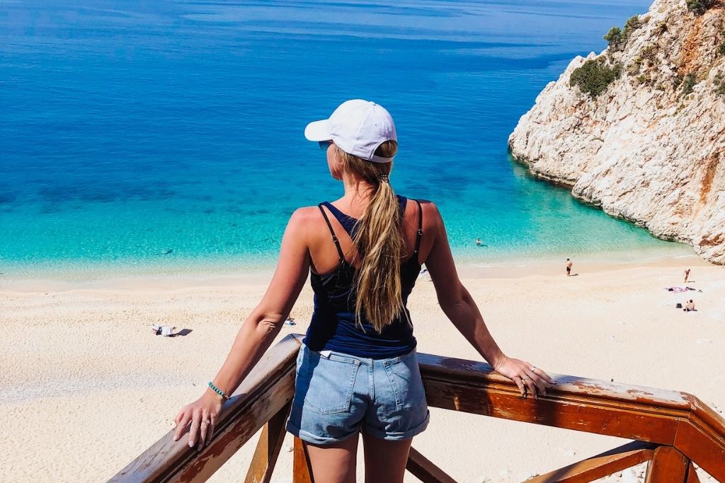 Kate wearing a white cap and denim shorts leans against a wooden railing, overlooking a stunning beach in Turkey with crystal-clear turquoise waters and rugged white cliffs. The sunny day highlights the serene and inviting coastal scenery.