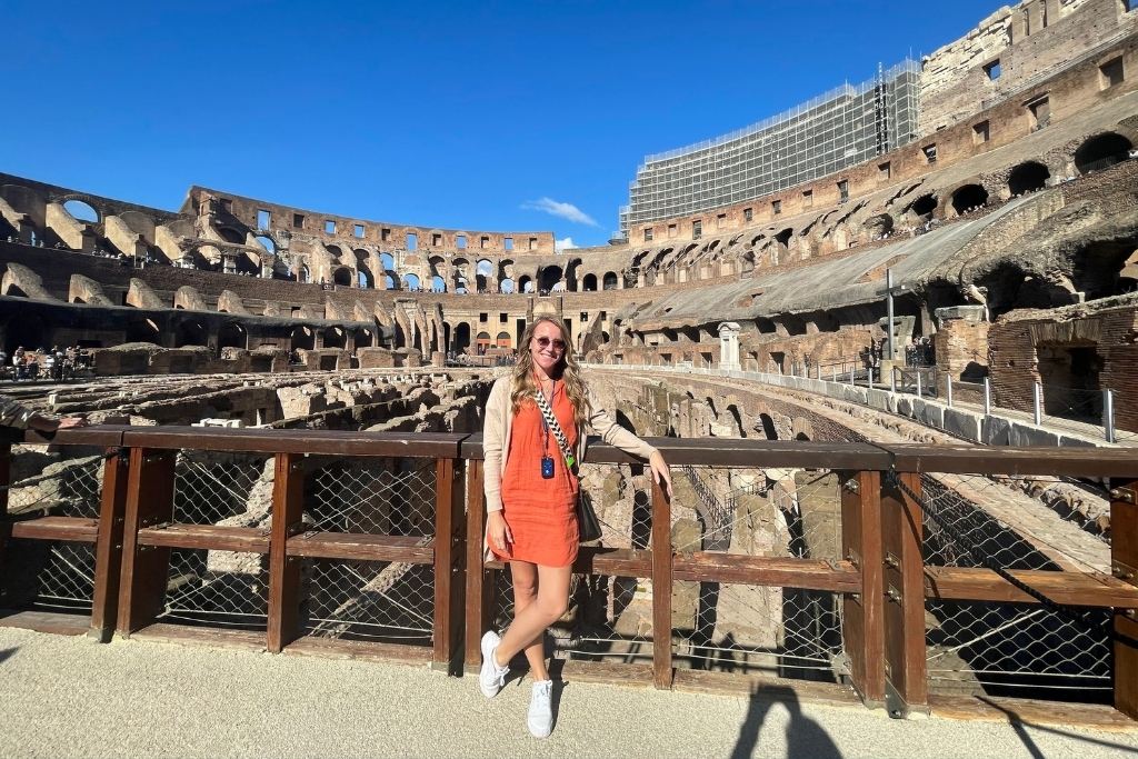 Kate from Kate's Crossing Blog standing inside the historic Colosseum in Rome, Italy, leaning against a wooden railing. She is wearing an orange dress, white sneakers, sunglasses, and a guide lanyard, smiling under the bright blue sky. The iconic ancient amphitheater's arches and stone structures provide a stunning and timeless backdrop.