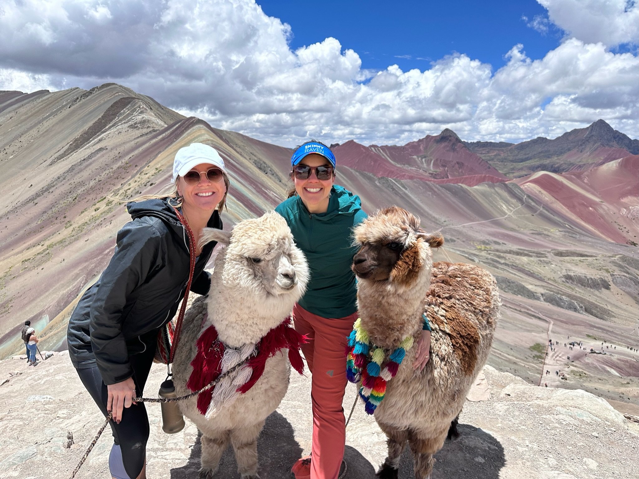 Kate and a friend posing with two alpacas adorned in colorful decorations at Rainbow Mountain in Peru. The vibrant, striped mountain landscape stretches behind them under a partly cloudy sky, highlighting the unique geological formations of the Andes. The scene captures the spirit of adventure and cultural connection in a stunning natural setting.