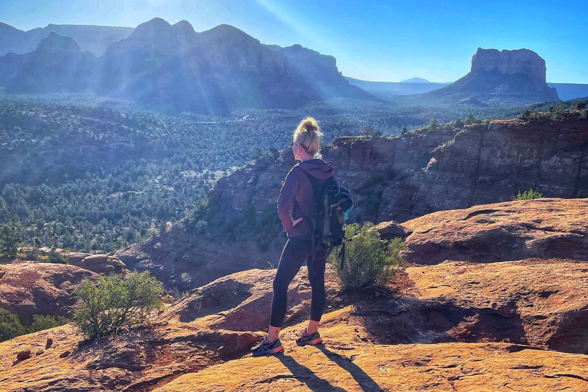 Kate stands on the rocky edge of Cathedral Rock in Sedona, gazing at the expansive valley below, with her backpack and hiking gear on, surrounded by stunning red rock formations under a clear blue sky.