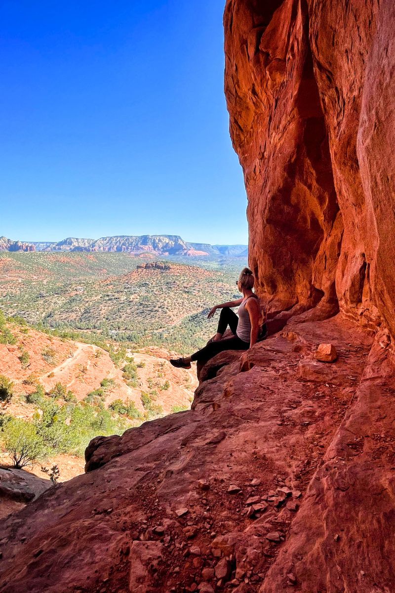 Kate sitting on a ledge with her back against the red rock formations of a cliff, gazing out at the expansive view of Sedona's landscape under a clear blue sky.