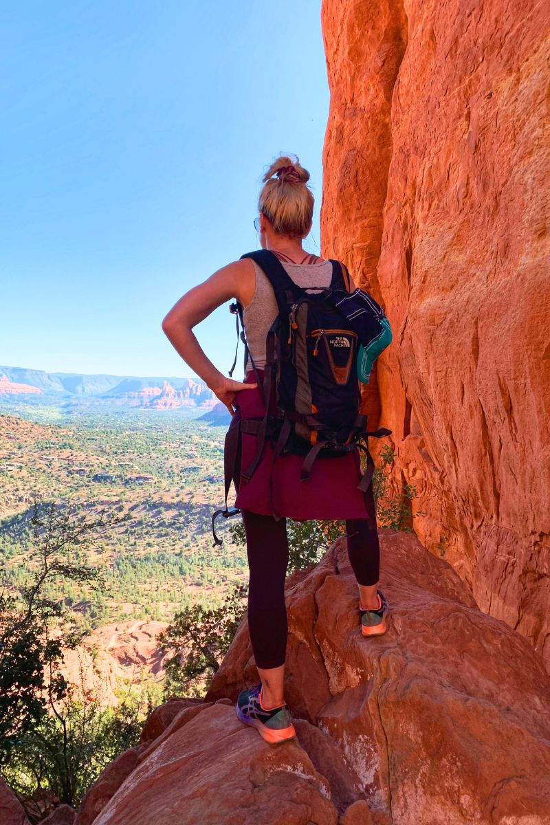 Kate standing on a rock ledge, admiring the expansive view of Sedona's landscape from Cathedral Rock, with her backpack on and hands on her hips.