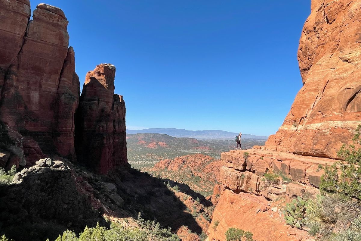 Kate stands on the edge of a cliff at Cathedral Rock in Sedona, surrounded by towering red rock formations and overlooking a vast, scenic valley under a clear blue sky.