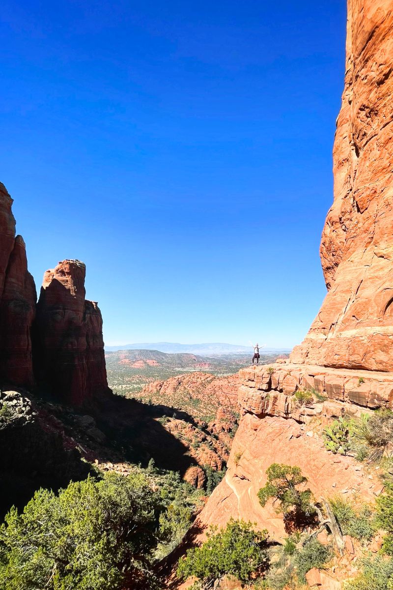 Kate stands with arms up on the edge of a cliff at Cathedral Rock in Sedona, with breathtaking views of the surrounding red rock formations and valley under a clear blue sky.