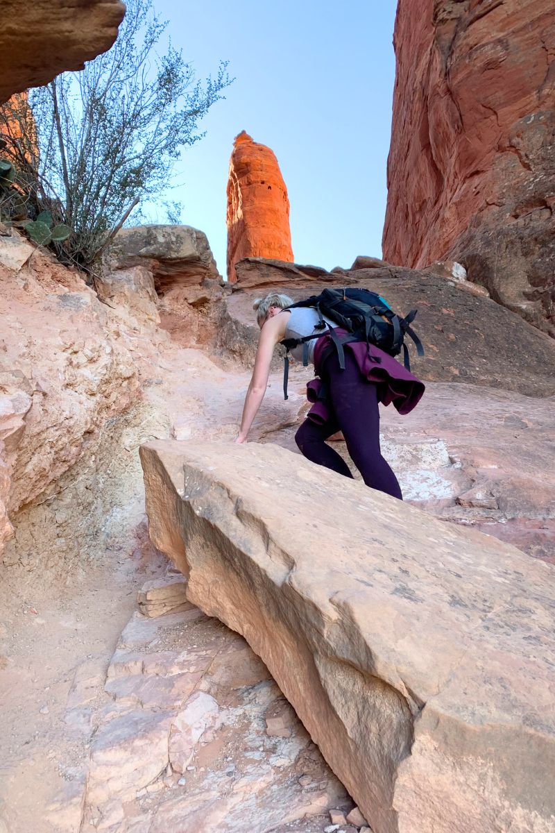 Kate climbs up a steep rocky trail to the needle spire, with the towering red rock spire in the background, showcasing the rugged beauty and challenge of the hike.