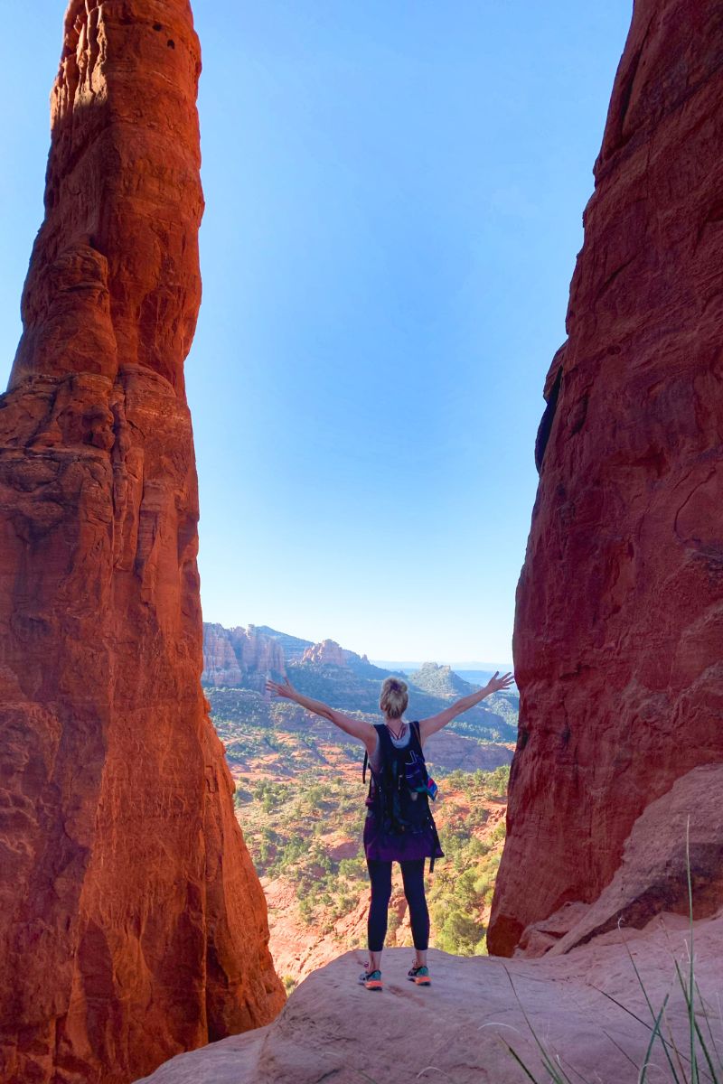 Kate stands with arms up between two towering red rock spires at Cathedral Rock in Sedona, arms outstretched, with a stunning view of the valley and distant formations.