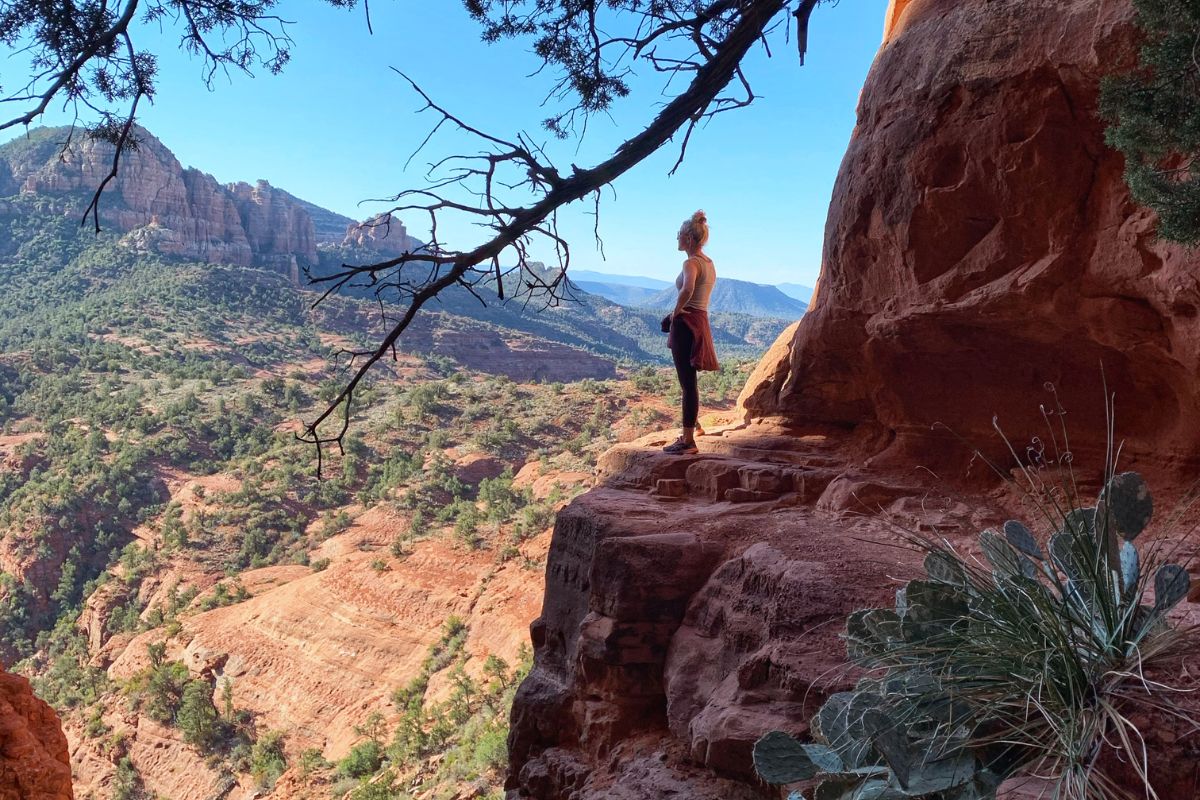 Kate stands on a rocky ledge on a secret cliff trail in Sedona, overlooking a vast expanse of red rock formations and greenery, capturing the beauty and tranquility of the landscape.