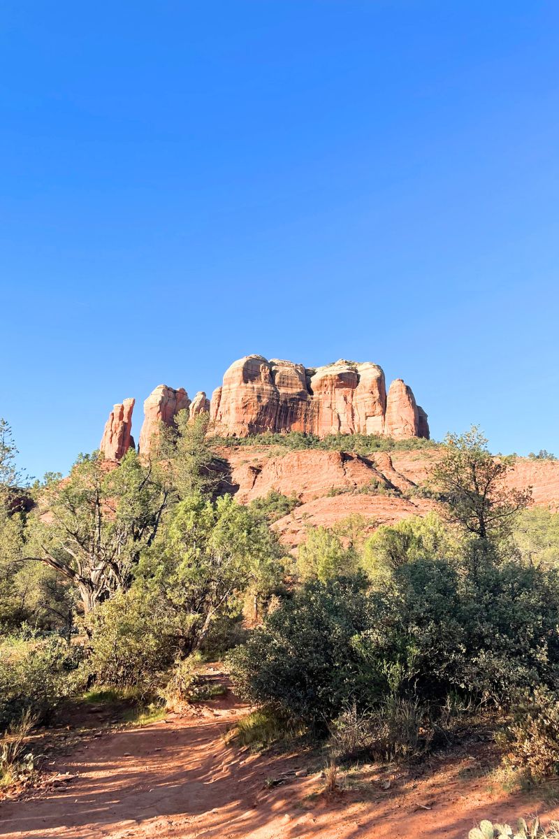 A scenic view of Cathedral Rock in Sedona, captured on a clear day with a dirt trail and bright green vegetation in the foreground.