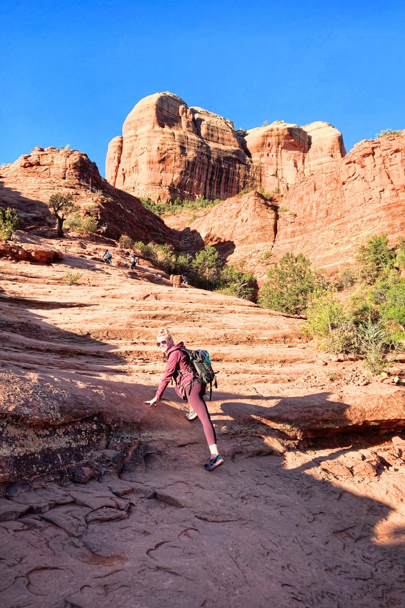 Kate, wearing a backpack and sunglasses, climbs up the rocky trail of Cathedral Rock in Sedona, surrounded by towering red rock formations and clear blue skies.