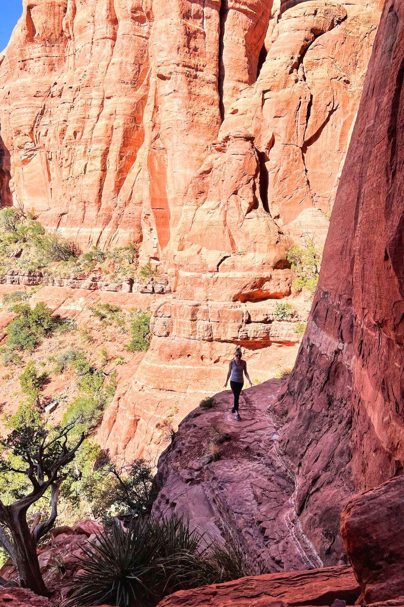 Kate hiking along a narrow trail on the red rock formations of Cathedral Rock in Sedona, with the majestic cliffs towering above her.