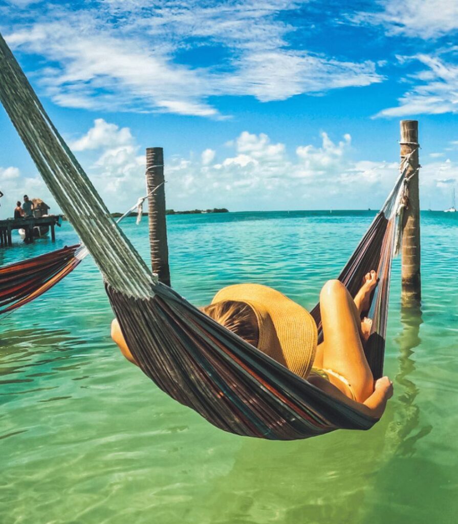 Kate relaxing in a hammock over turquoise waters at Caye Caulker, Belize, wearing a wide-brimmed hat under a vibrant blue sky.