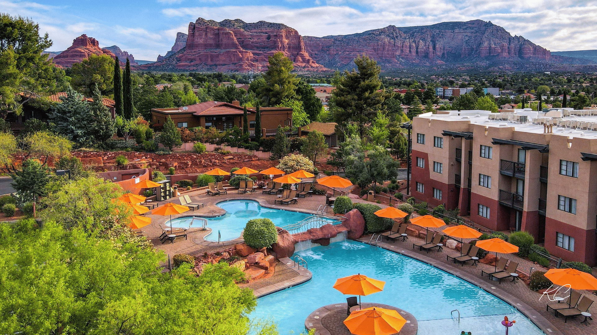 A resort in Sedona with a stunning pool area featuring orange umbrellas, lush greenery, and a backdrop of red rock formations under a partly cloudy sky.