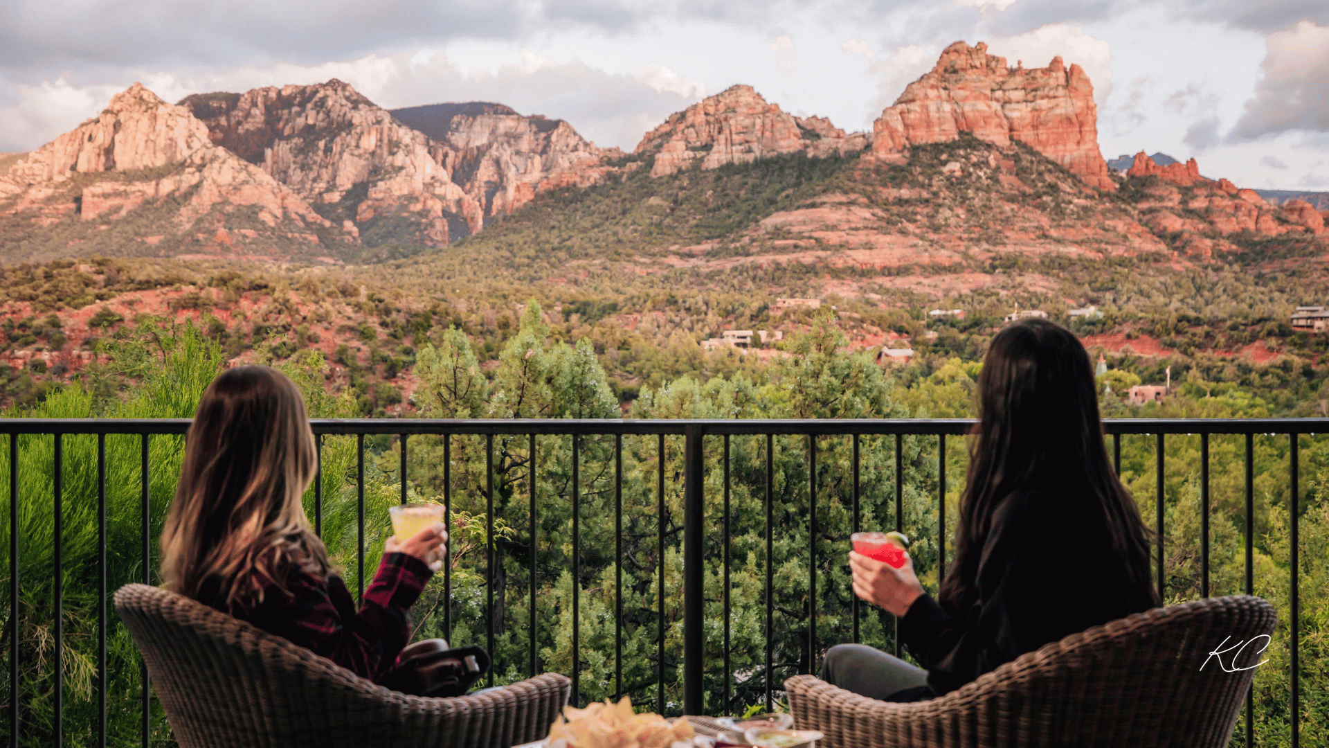 Two women enjoying drinks on a balcony with a stunning view of Sedona's red rock formations and desert landscape in the background.