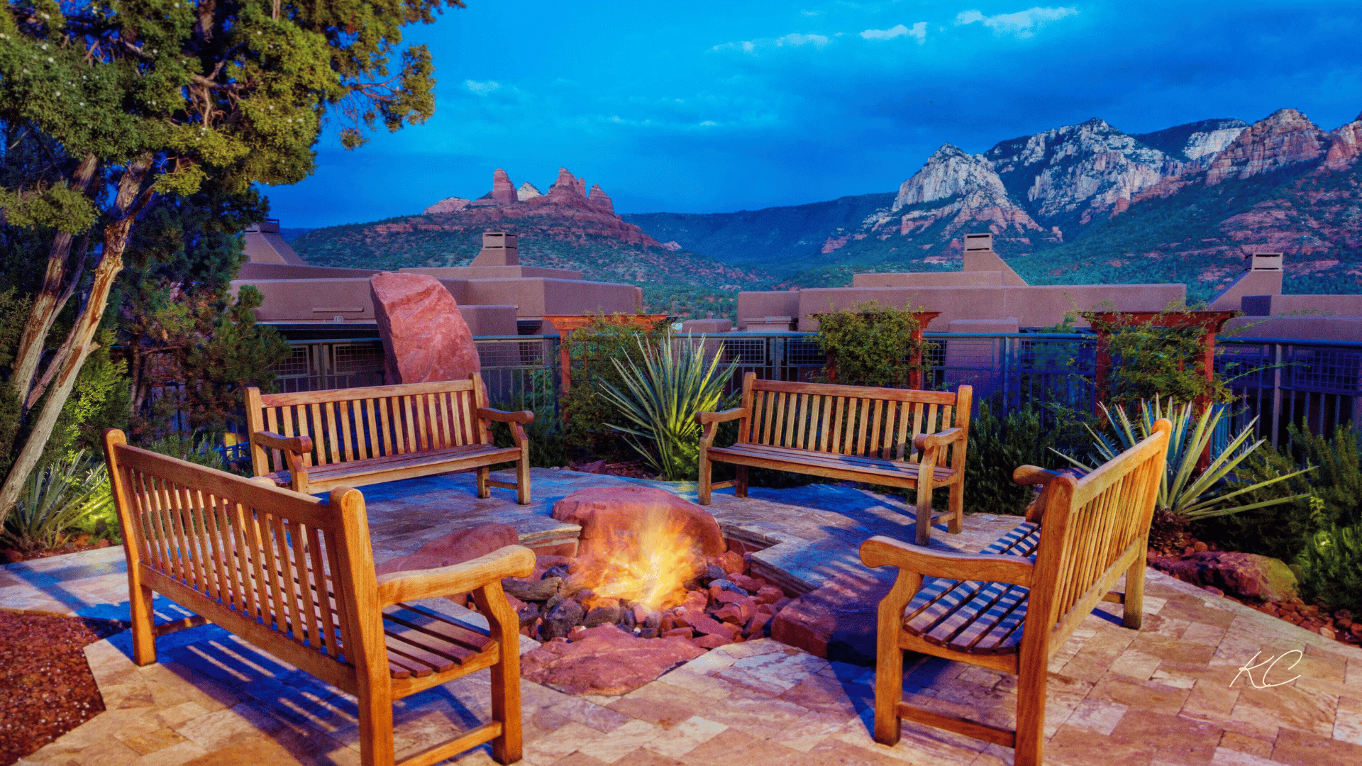 A cozy outdoor fire pit surrounded by wooden benches, with a stunning view of Sedona's red rock formations and mountains under a twilight sky.