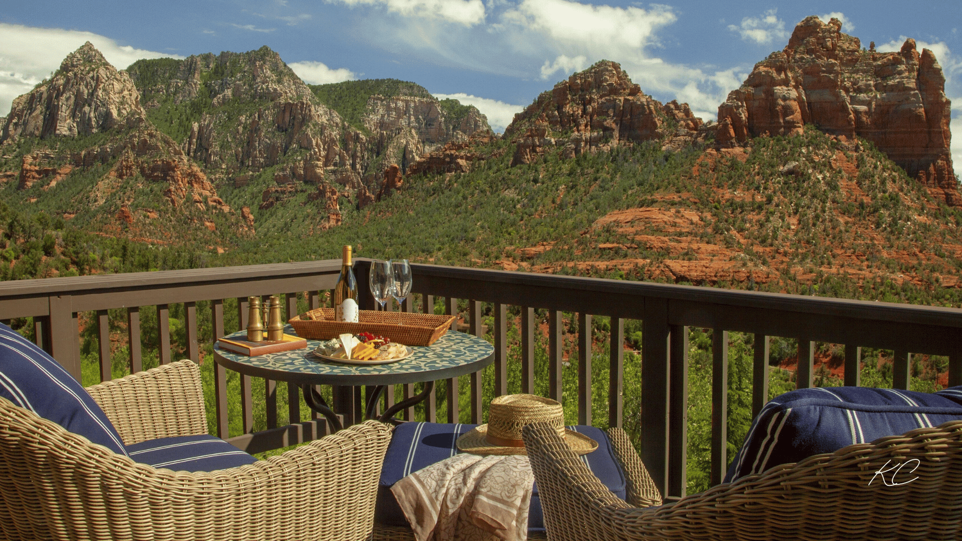 A cozy balcony with wicker chairs, a table set with wine and snacks, overlooking a stunning view of Sedona's red rock formations under a bright blue sky.