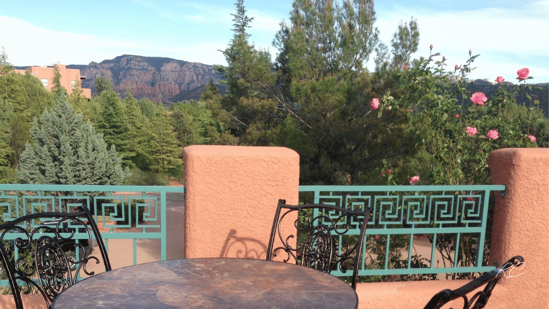 A charming balcony with wrought-iron chairs and a table, surrounded by lush greenery and pink flowers, offering a scenic view of Sedona's mountains.