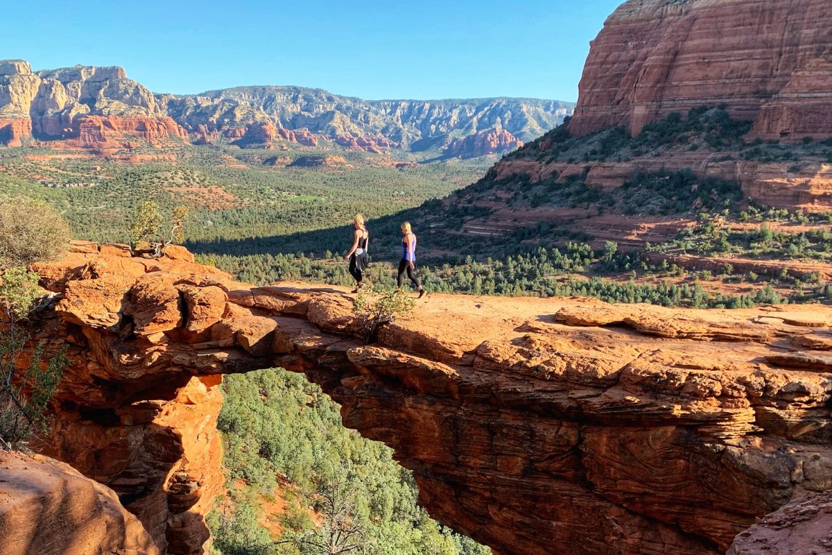 Kate and a companion walking across the stunning natural sandstone arch of Devils Bridge, surrounded by Sedona's breathtaking red rock formations and lush green valleys during the Devils Bridge Hike.