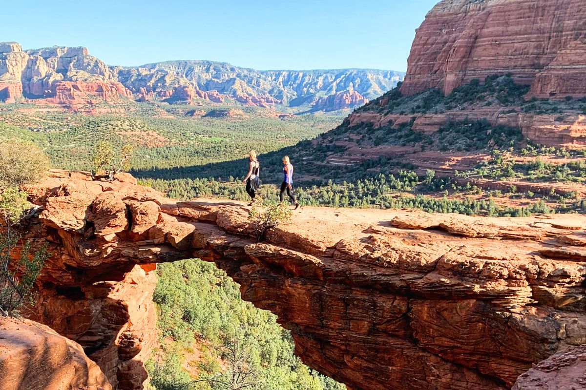 Two hikers, including Kate, walking across the natural red rock arch of Devil's Bridge with a stunning backdrop of Sedona's red rock formations and green valleys.