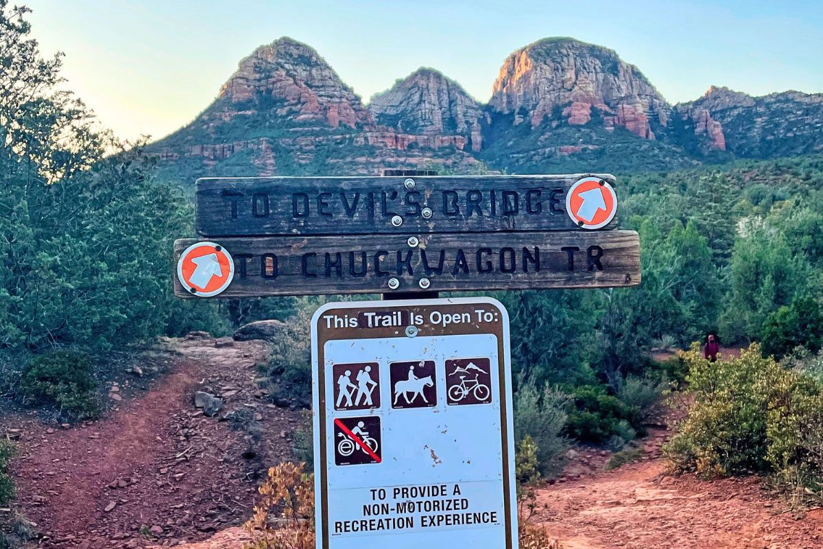 Trail sign pointing to Devil's Bridge and Chuck Wagon Trail in Sedona, Arizona, with a scenic view of the red rock formations in the background.