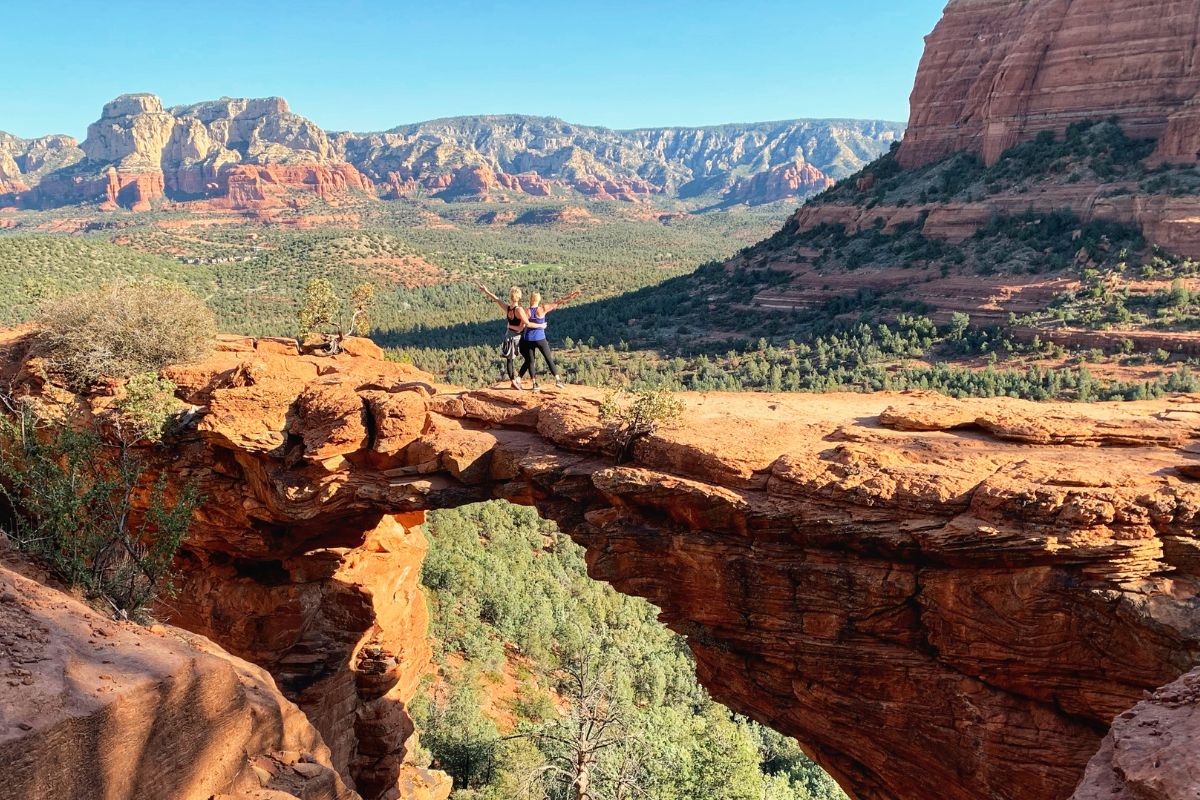 Kate and a friend standing on the natural sandstone arch of Devils Bridge, surrounded by Sedona's stunning red rock formations and expansive forested valleys during the Devils Bridge Hike.