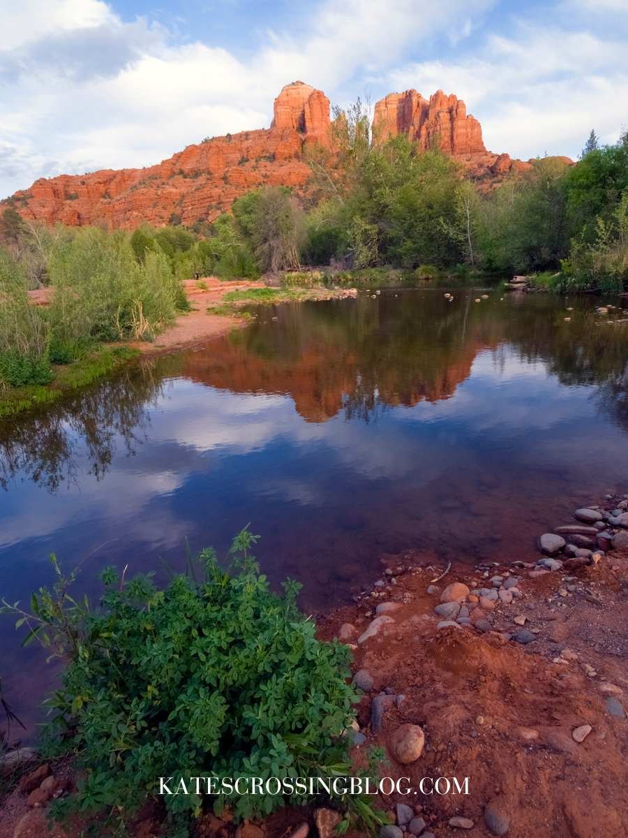 Picture of Cathedral rock in the back ground and its mirroed image in the calm water puddles of Oak Creek at Red Rock Crossing Trail. There are some clouds in the sky but it is still sunny.