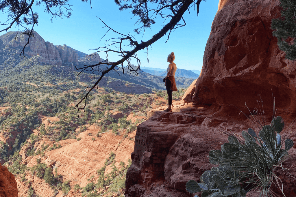 Kate standing on a cliff ledge surround by cacti, looking out at the desert views.