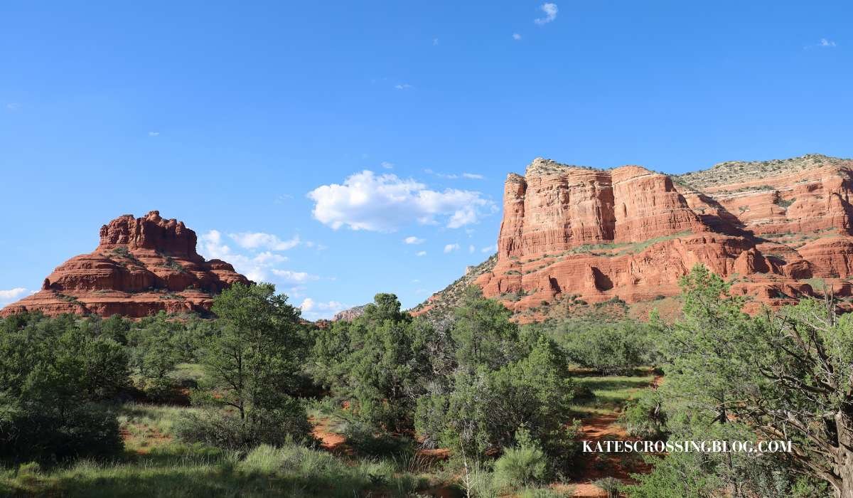 Picture of Bell Rock and Courthouse Butte which are two red rock formations that are right next to each other making this one of the best hikes in Sedona. There are green juniper trees below these towering rock formations with a blue bird sky above.