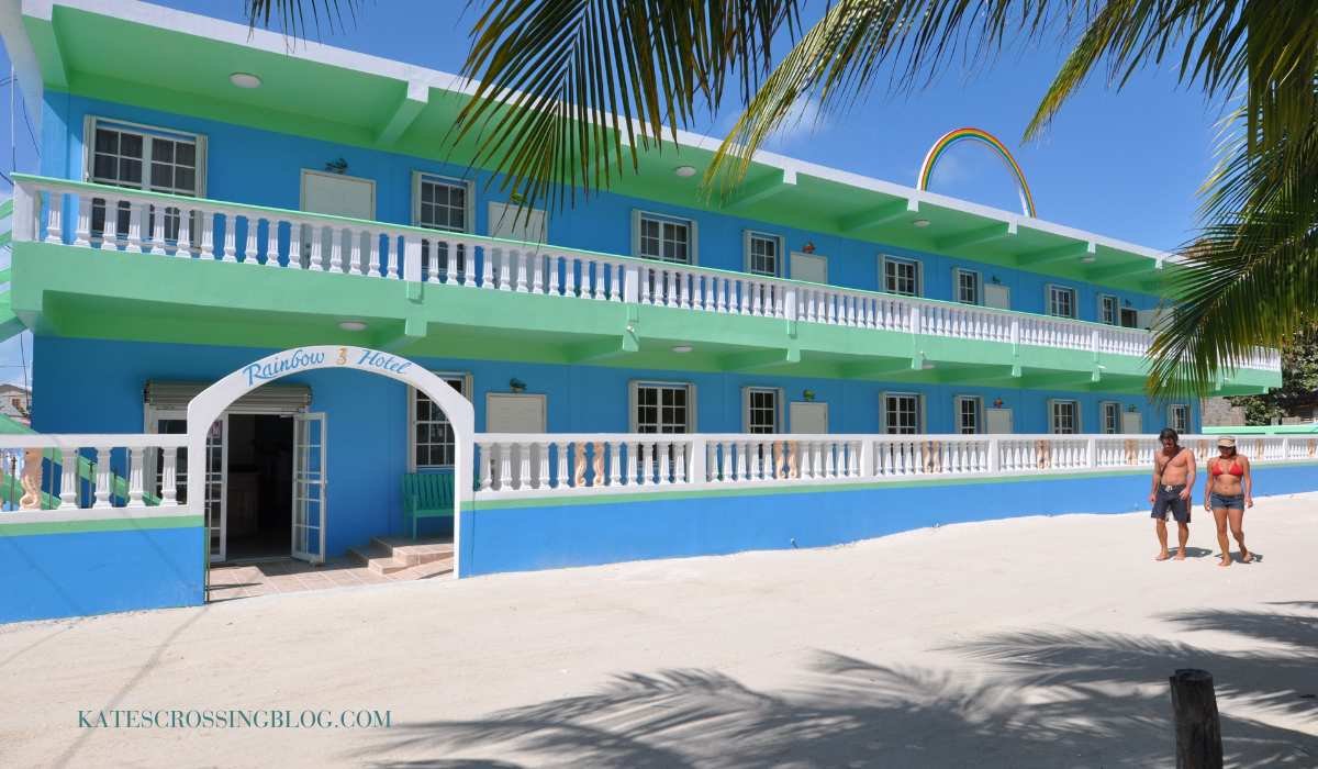 front view of Rainbow Hotel. Its two stories tall, painted in a baby blue and teal green with white trim. The street out front is white beach sand and two people are walking by wearing swimsuits.