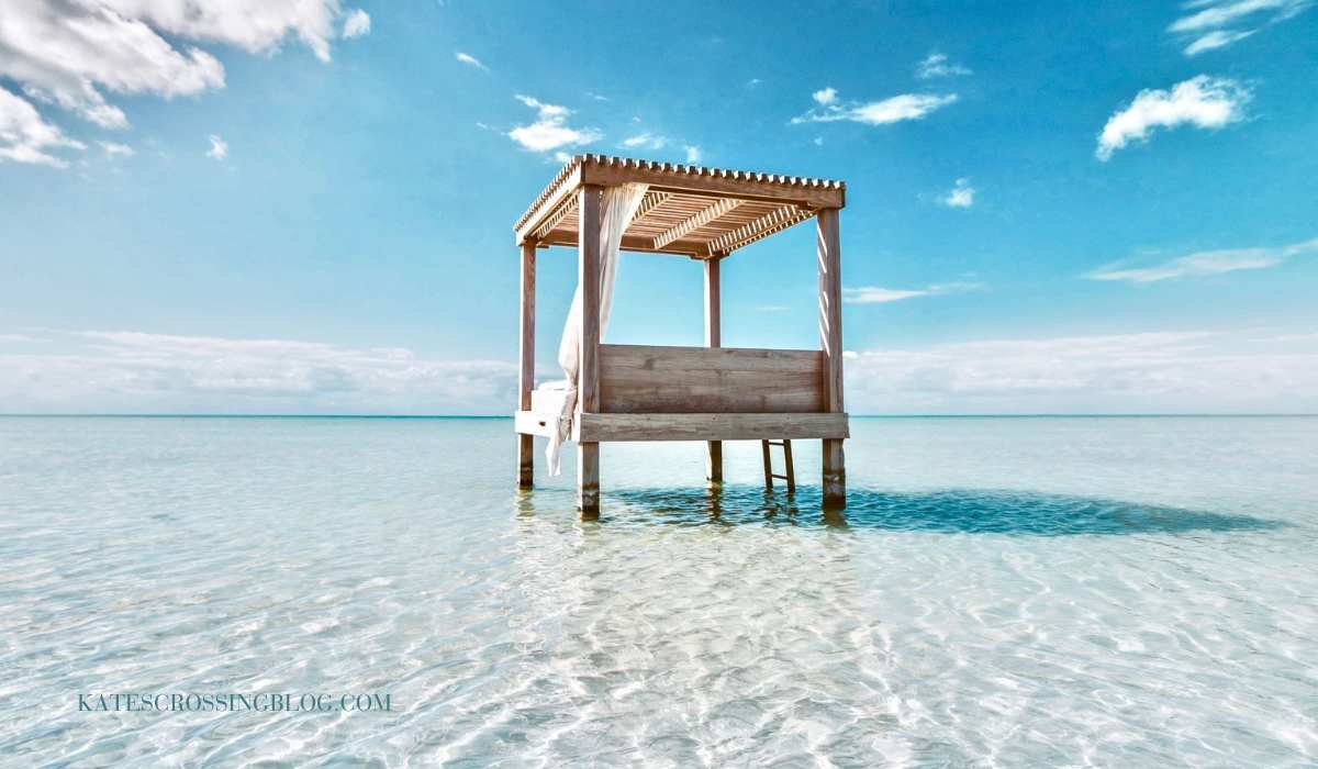 A private beach cabana out in the water of crystal clear turquoise waters and white sand. The sky is bright blue. There is a ladder you climb to get up into the cabana that has a mattress to lounge on.