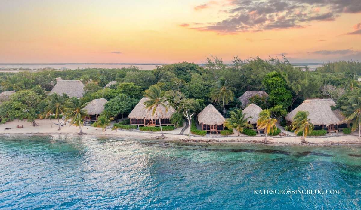arial view of Turtle Inn cabana's along a beach, surrounded  by jungle vegetation and palm trees. The water along the beach is turquoise and the ski is a mix of light yellow and pinks as the sun sets.
