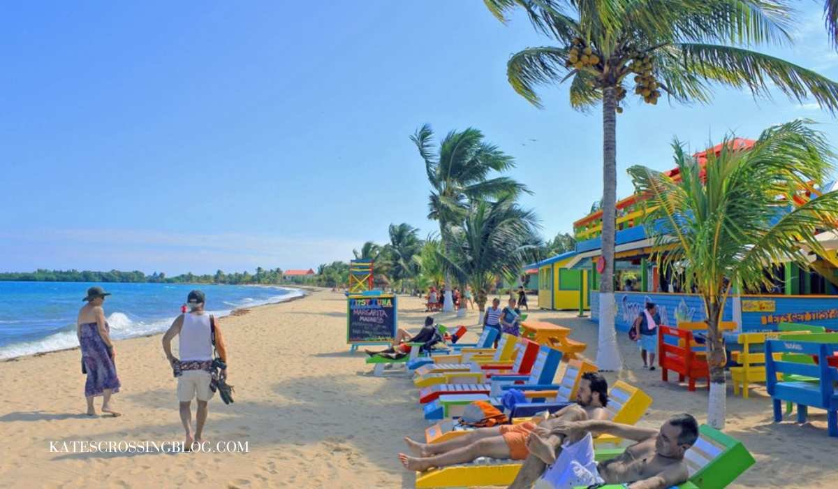 A beach in front of Placencia Villas, lined with rainbow colored beach lounge chairs. The picnic tables and restaurant is also painted in rainbow colors. There are palm trees all along the shore and people enjoying the sunny beach.