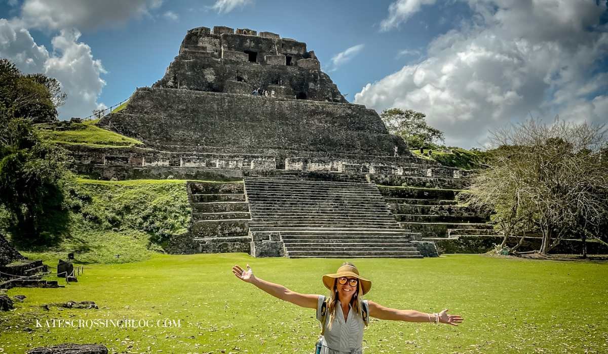 Picture of me standing in front of the main temple in Xunantunich. There is no one else there. The temple has stone steps leading up the front with perfectly manicured greeen grass all around the base. The day is warm and sunny. I'm wearing a dress.