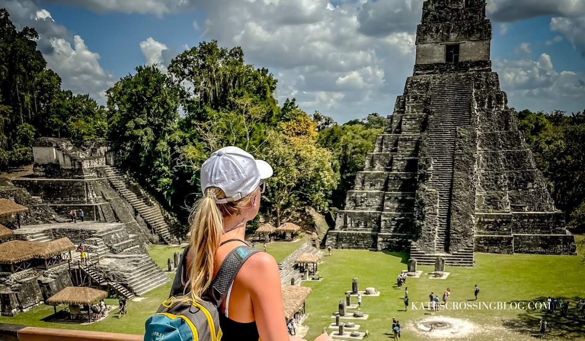 Picture of me gazing up at the most popular mayan temple in Tikal. The towering stone structure has stair teps almost to the top. the grass is perfectly manicured around the base. The dense jungle is in the background. It's a sunny day.