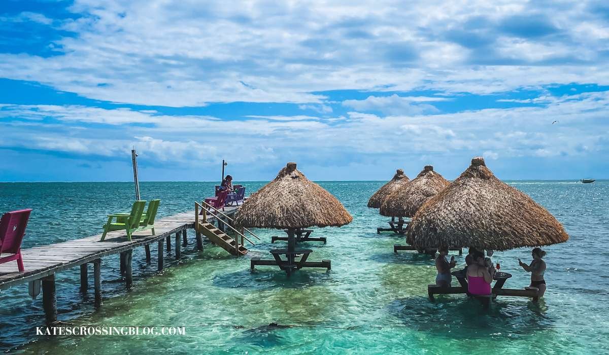 picture of picnic tables covered by thatched roofs out in the sallow turquoise waters off the shore of Caye Caulker. People are wading in the water with a drink in hand enjoying the sunshine.