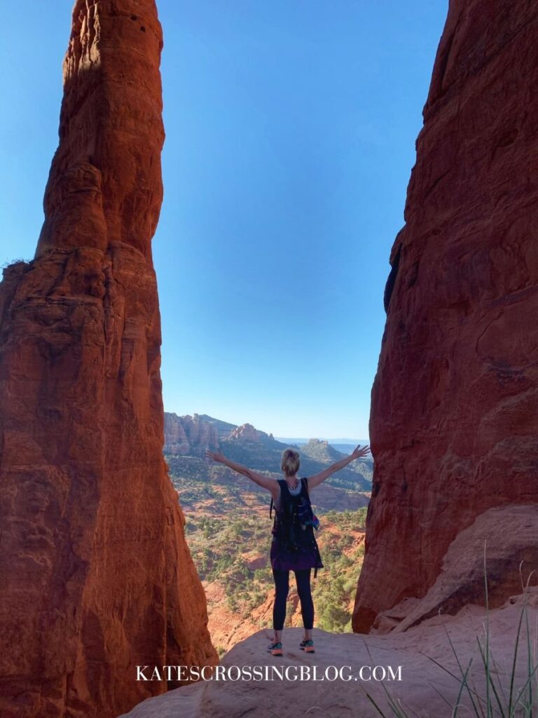 Kate standing triumphantly between two towering red rock formations at Cathedral Rock in Sedona, Arizona, with arms outstretched and a stunning vista of red rock landscape and blue sky in the background.